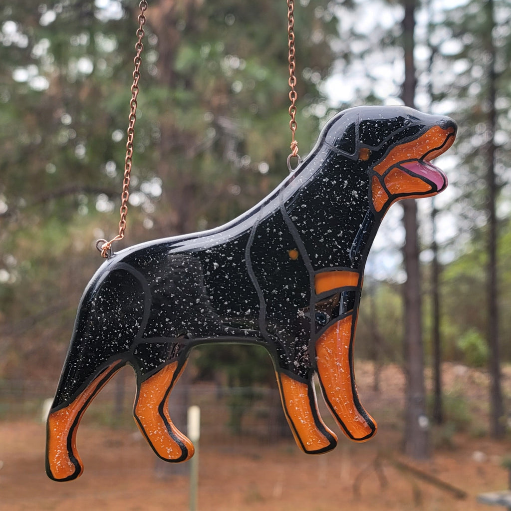 Decorative glass Rottweiler dog figure hanging outdoors with trees in the background Rottweiler Glass Urn Ashes-Infused-Glass Ornament   A glass cremation urn suncatcher with a silhouette of a Rottweiler dog with cremation ashes inside. infusion glass artist Joele Williams ashes infused glass