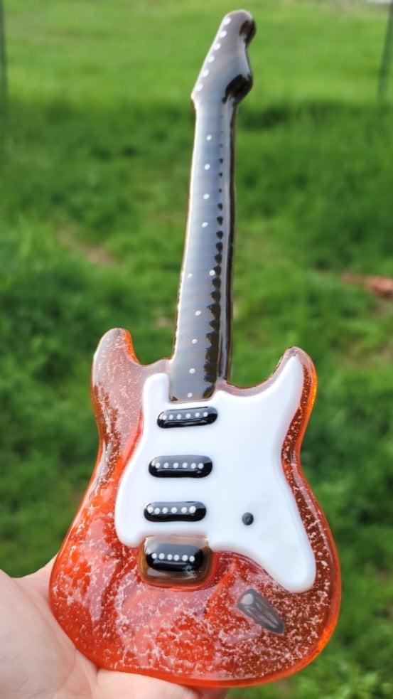 hand crafted Guitar made from glass with Cremation Ash Infused inside Glass Memorial Keepsake  handmade by artist joele williams infusion glass ashes infused glass. A decorative guitar-shaped ashes infused glass miniature guitar replica  The guitar is held in a hand against a blurred grass background. A 10 inch miniature glass electric guitar with a red and white design, featuring black strings and a black pickguard.
